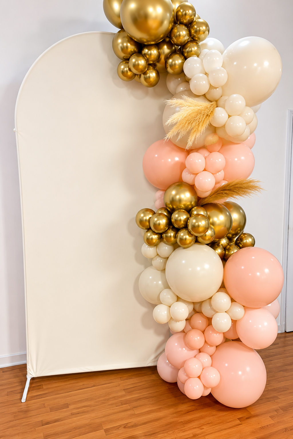 Decorative balloon arch with gold, cream, and blush pink balloons, positioned next to a white backdrop.