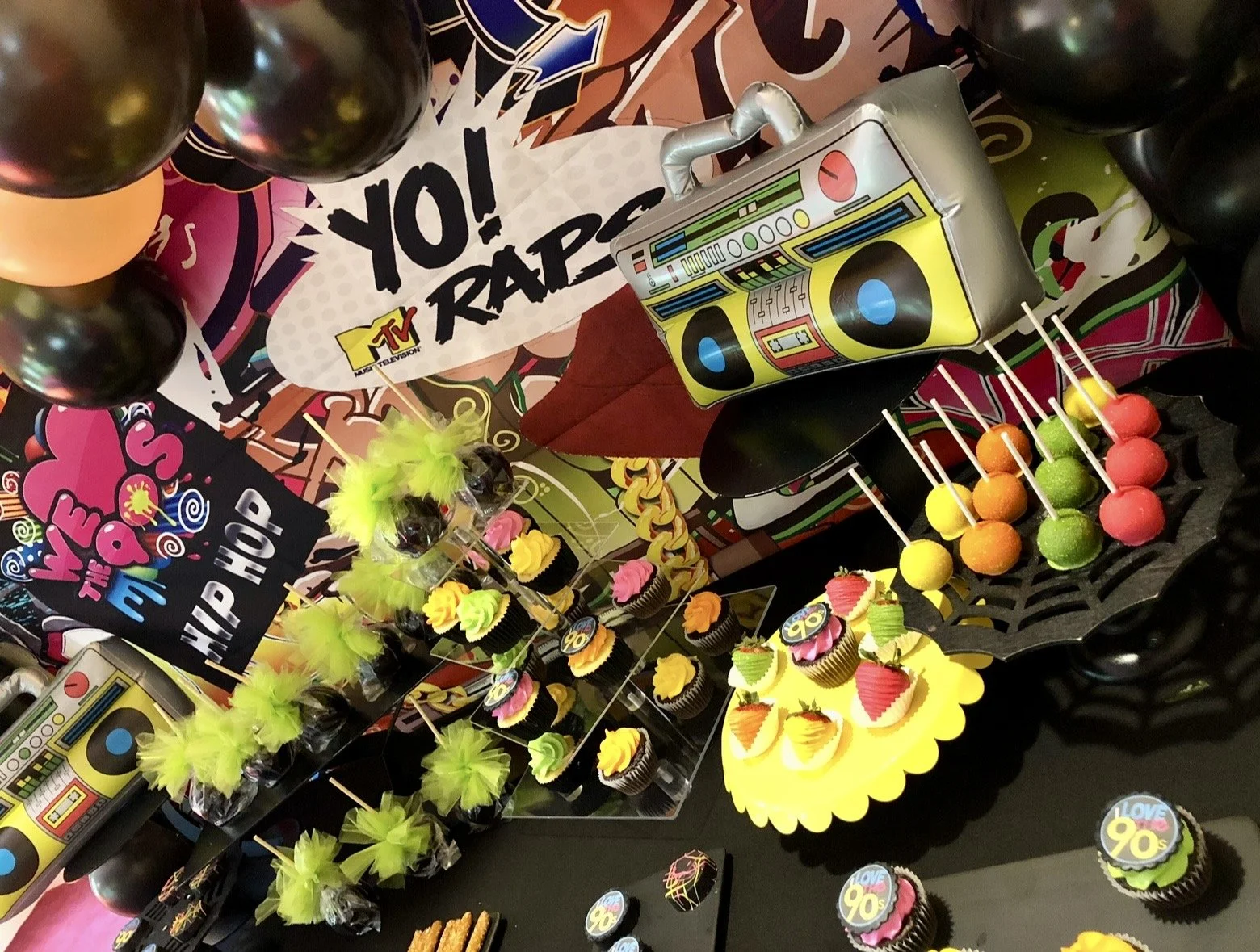 Colorful 90s-themed dessert table including themed cupcakes, lollipops, balloons, and a box with a boombox design.