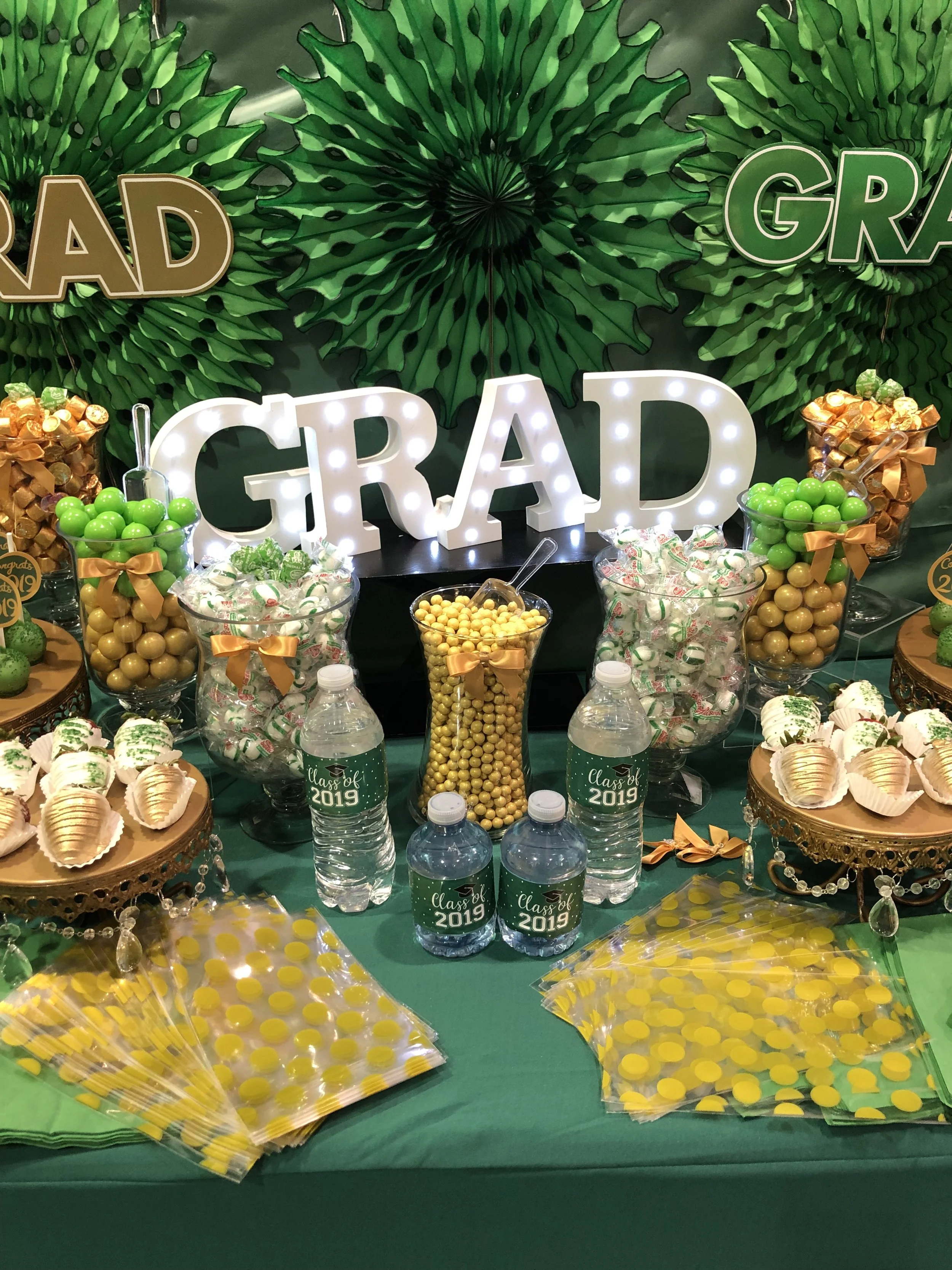 Decorative table with patriotic colors, celebrating a graduation in 2019. The table has green candies, cupcakes, water bottles with 'Class of 2019' labels, and a large illuminated 'GRAD' sign. The background features green foliage and partially visib