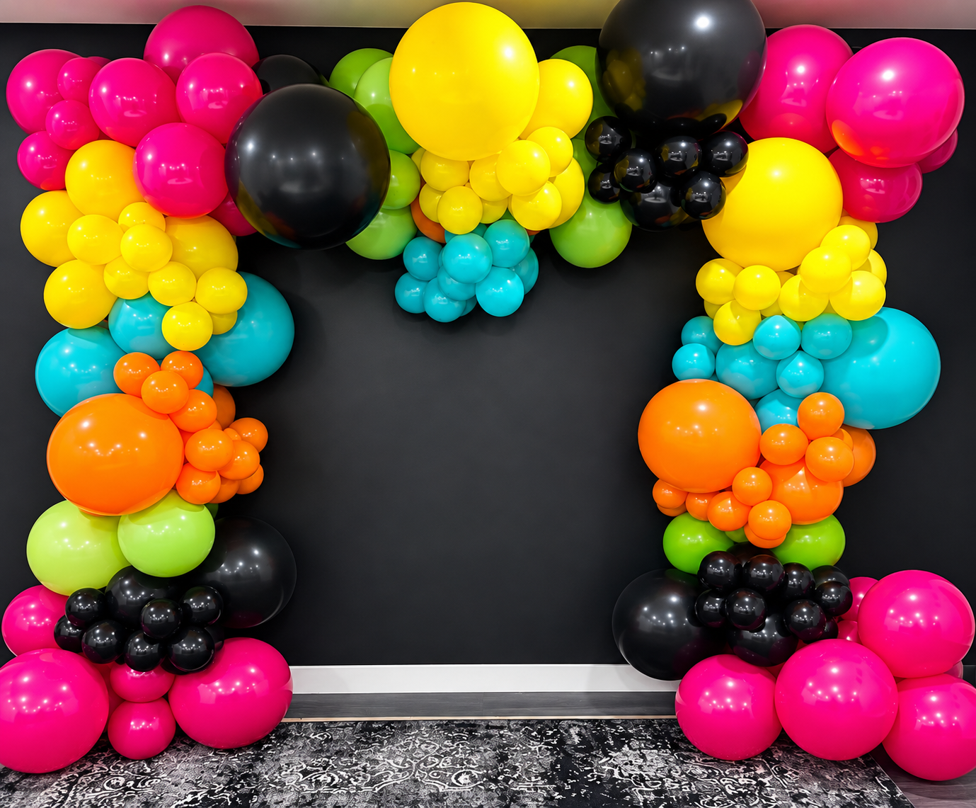 Colorful balloon arch with pink, yellow, green, blue, orange, black balloons arranged in a decorative pattern against a black wall.