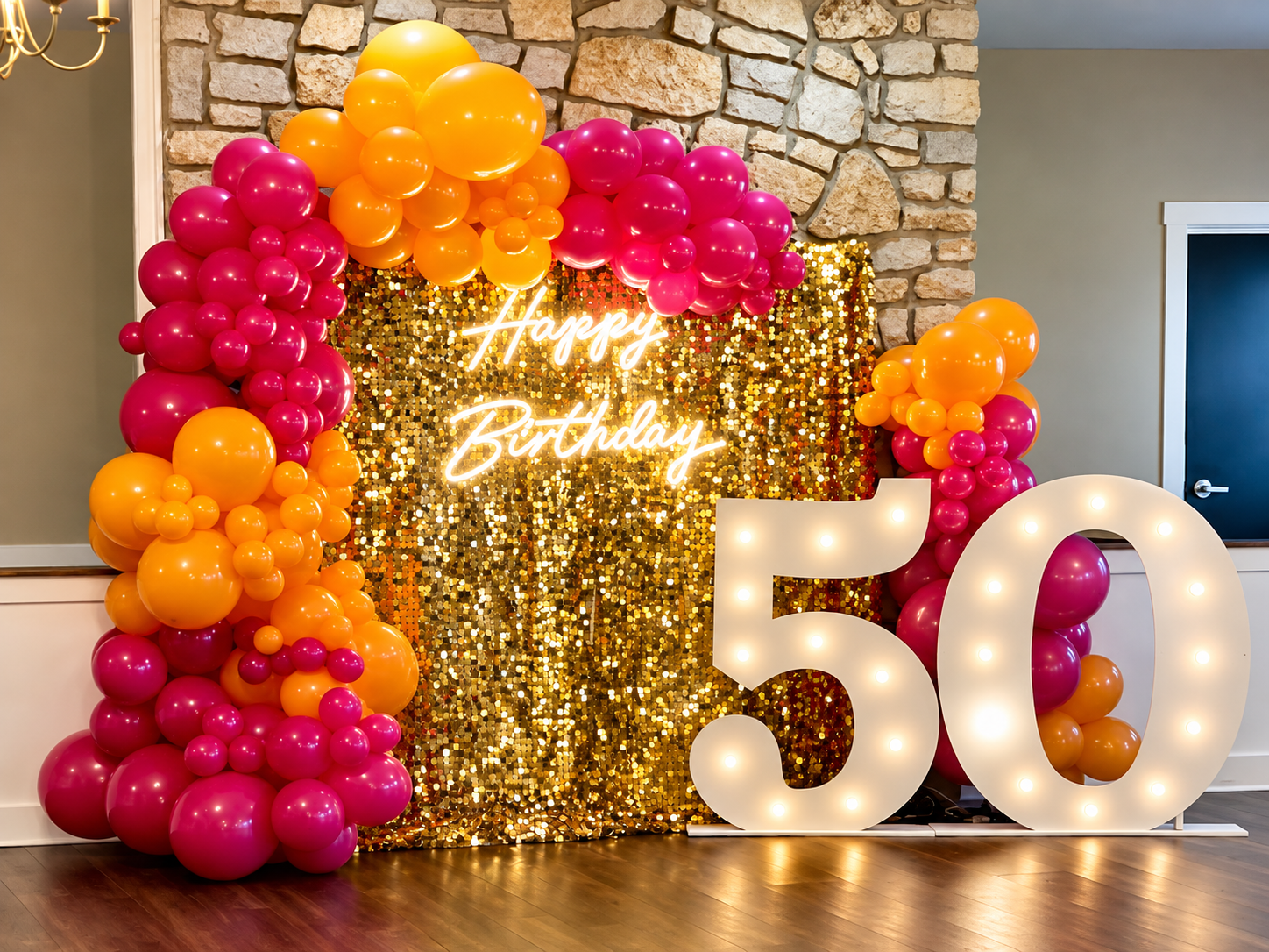 Decorative birthday celebration setup with a large illuminated '50', surrounded by pink and orange balloons, a sequined gold backdrop, and a neon sign that reads 'Happy Birthday'.