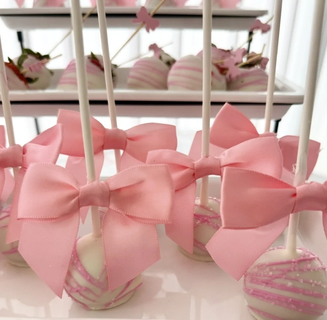 Pink and white chocolate-covered cake pops decorated with pink bows and pink drizzle, displayed on a tiered serving tray.