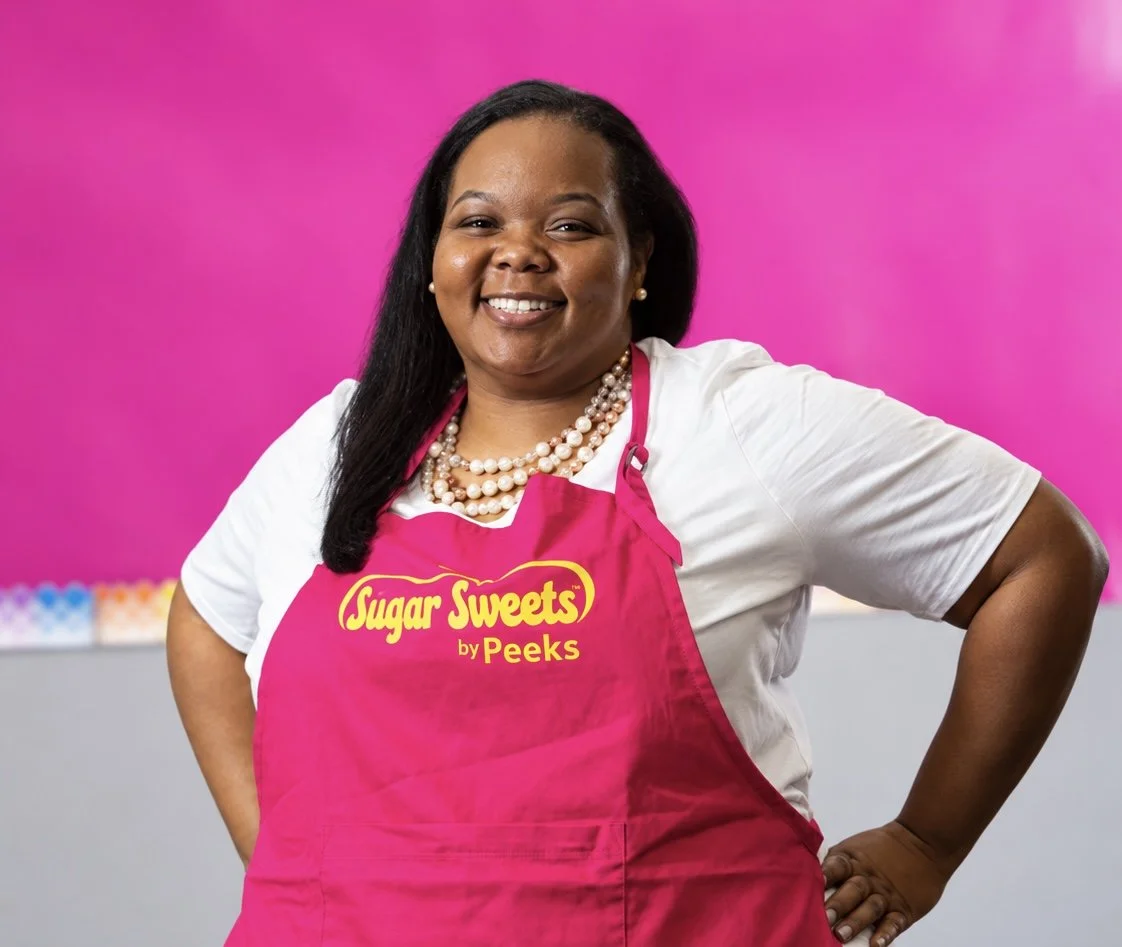 A woman smiling, wearing a white shirt, pearl necklace, earrings, and a pink apron with 'Sugar Sweets by Peeks' logo, standing in front of a vibrant pink background.