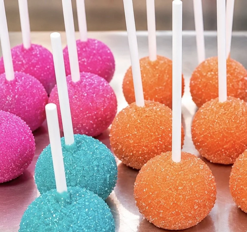 Colorful cake pops with sprinkles, arranged in pink, blue, and orange, each with a white stick.