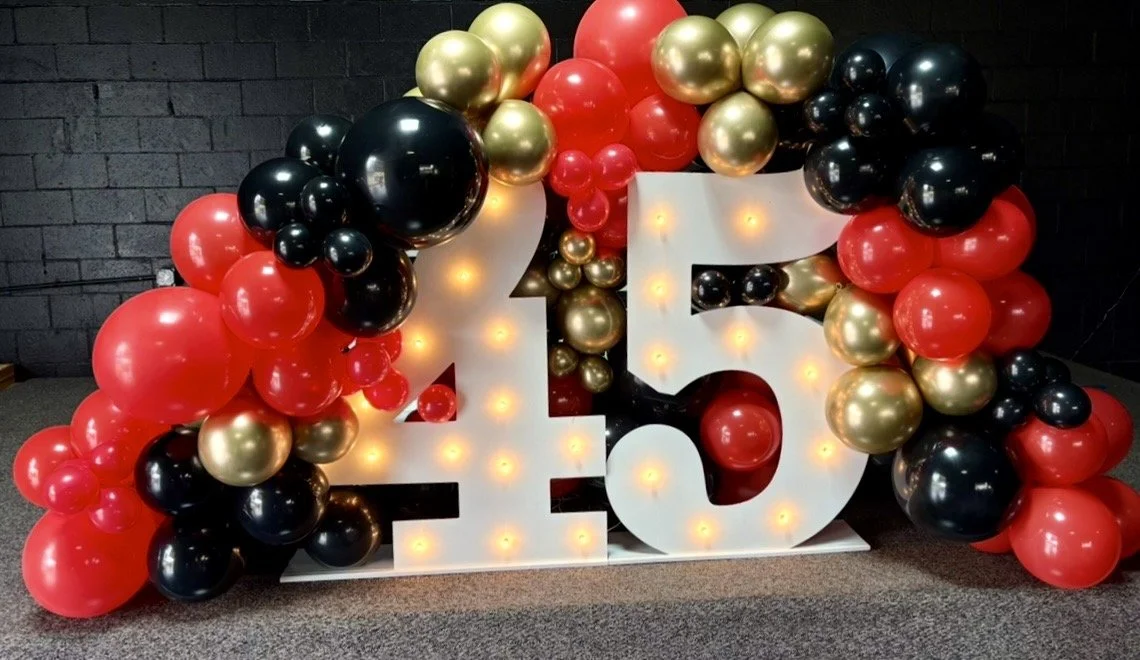 Decorative display featuring large illuminated white number 45 surrounded by red, black, and gold balloons for milestone birthday event by Sugar Sweets by Peeks. 