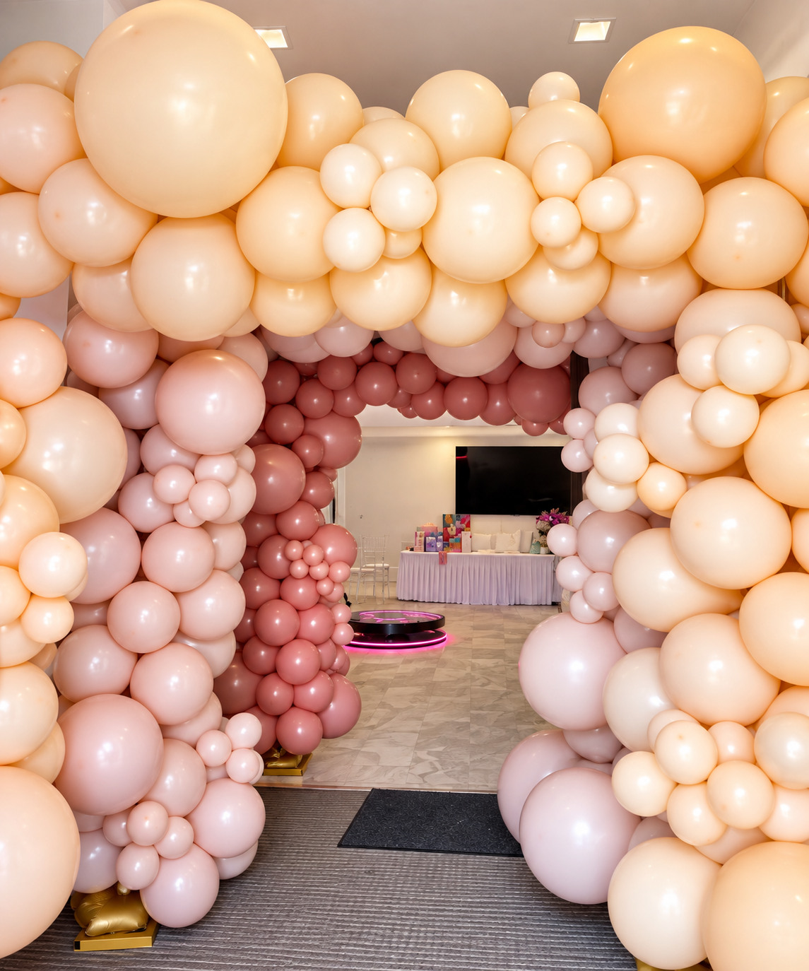Baby shower balloon arch with cream, pink, and blush balloons at an indoor celebration space, with a table of gifts and decorations in the background.