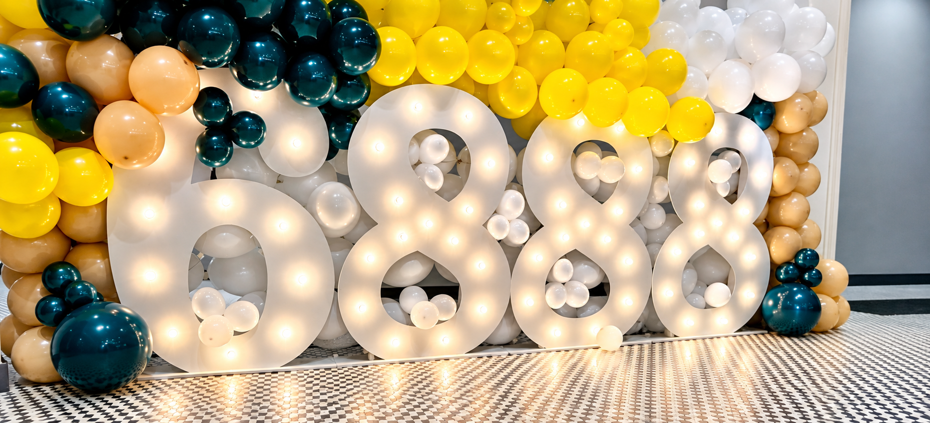 Large illuminated number display surrounded by colorful balloons in black, yellow, white, and beige, arranged for celebration by Sugar Sweets by Peeks.