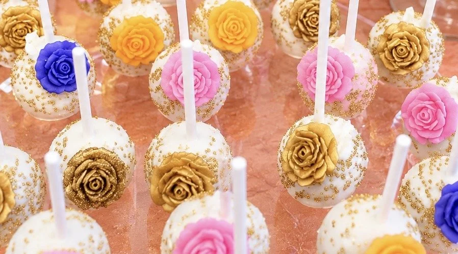 Cake pops decorated with white icing and gold, pink, purple, and yellow rose-shaped fondant toppers, arranged on a pink surface.