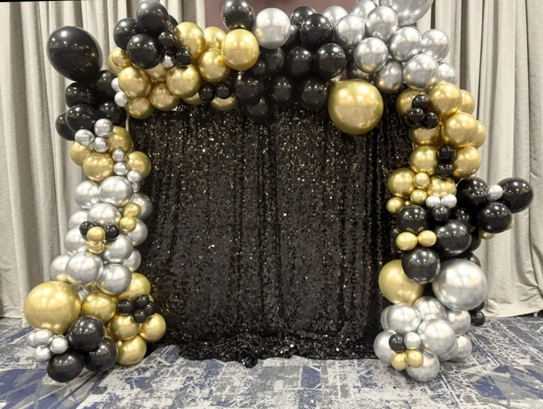 A black, shiny, sequin-covered backdrop with an arch of black, gold, silver, and white balloons in front of it, set against light-colored curtains and a patterned floor by Sugar Sweets by Peeks.