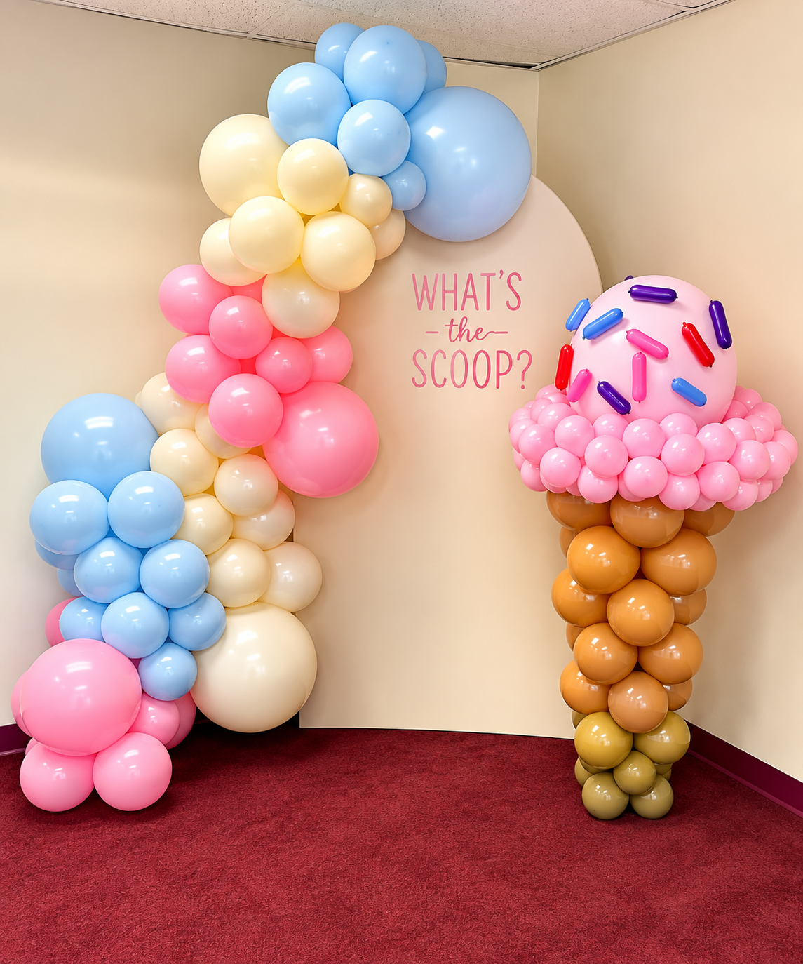 Decorative balloon arrangements resembling ice cream cones with pink, blue, cream, and brown balloons, and a sign that reads "What's the scoop?" on a beige backdrop by Sugar Sweets by Peeks.