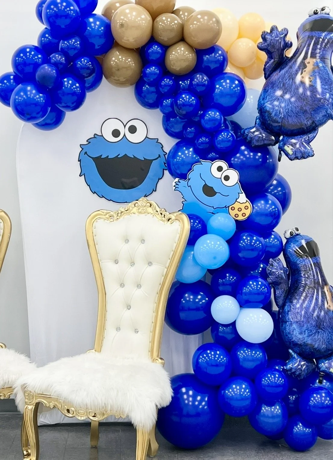 Decorative backdrop featuring blue and beige balloons, Sesame Street emoji cutouts of Cookie Monster with a cookie and another with a bowtie, and plush cookie monster balloons. A gold ornate chair with white tufted cushion and fur throw is placed in 
