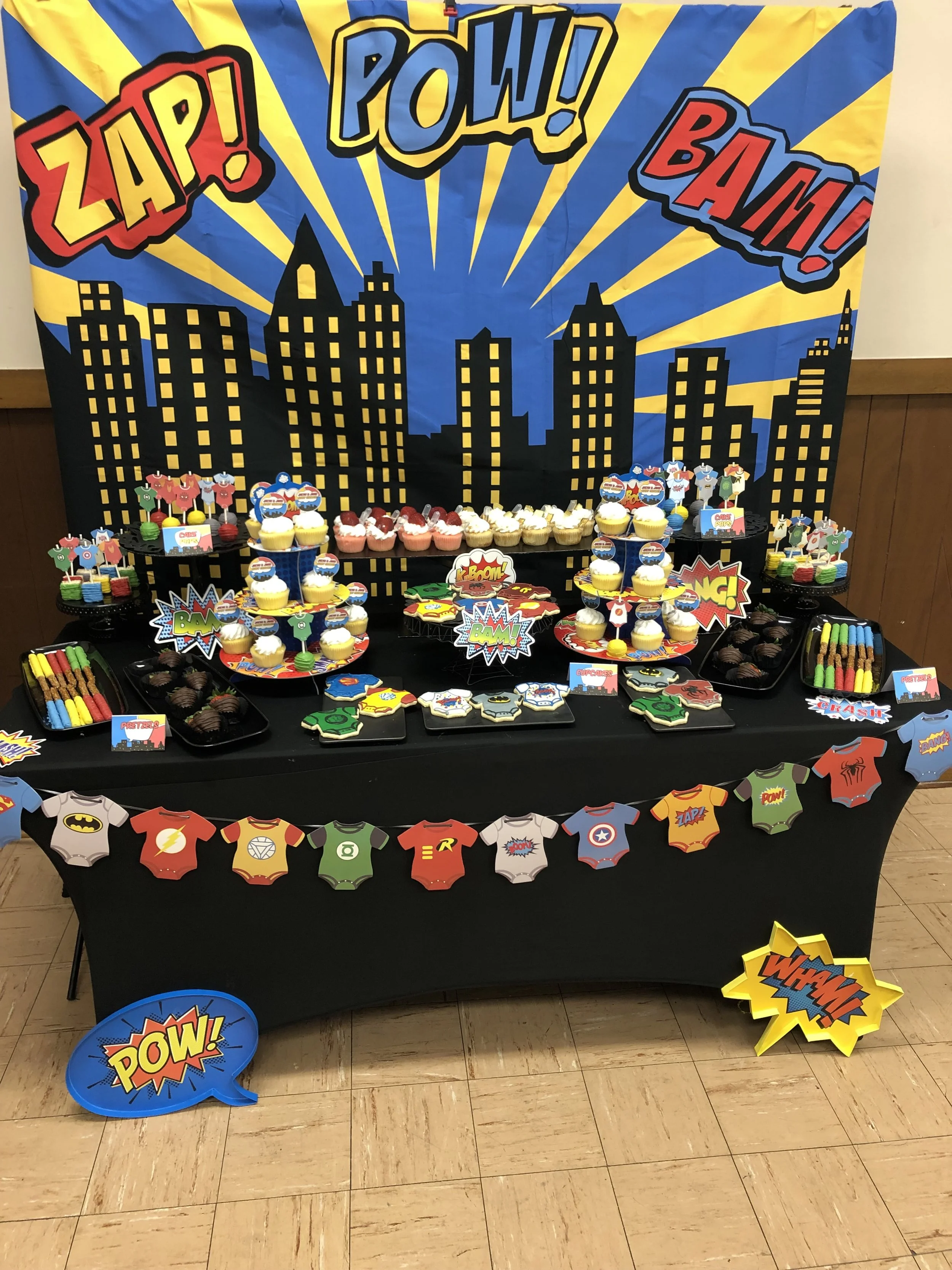 Superhero-themed dessert table with a city skyline backdrop featuring comic book style words The table has cupcakes, cookies, and candies decorated with superhero symbols, and colorful photo props and superhero speech bubbles.