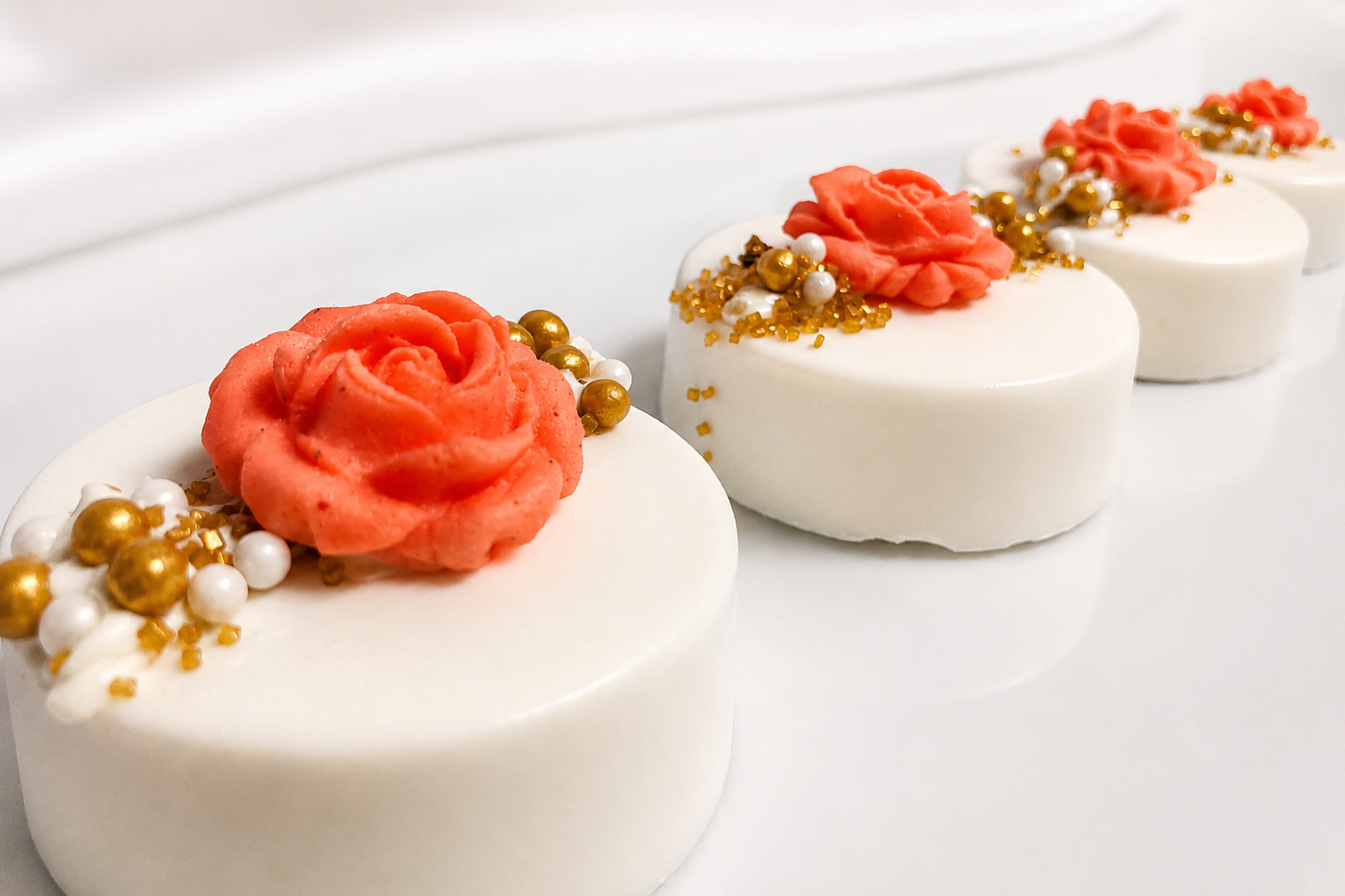 Line of small, round, white, and gold decorated cakes with pink flower-shaped frosting on top and gold, white, and pink sprinkles.
