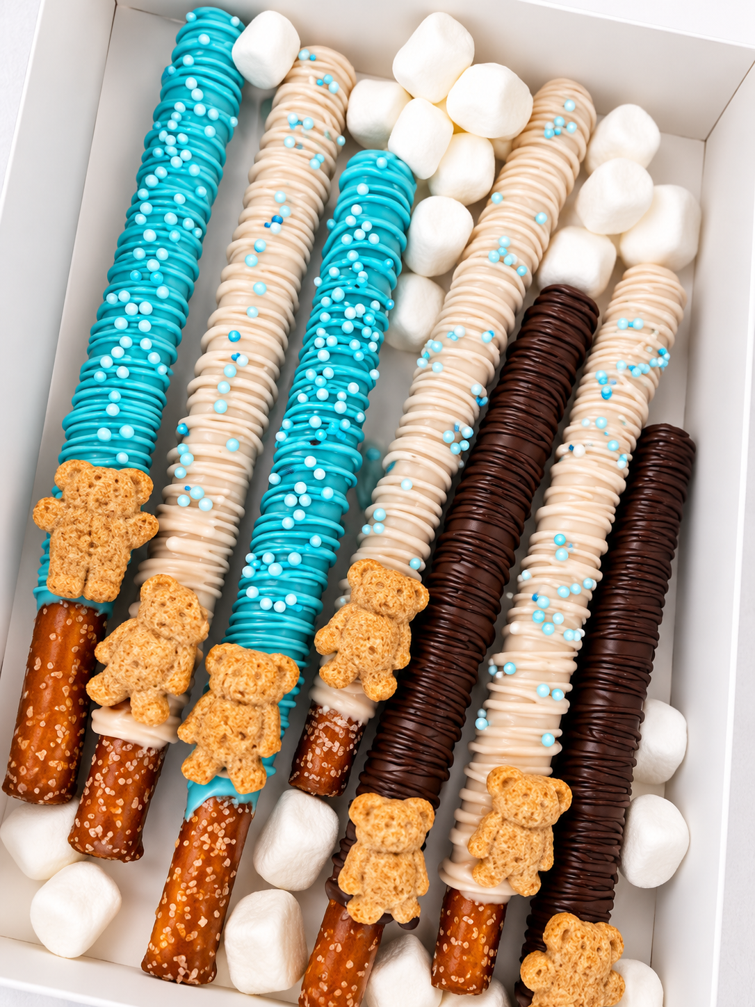 Box of decorated chocolate covered pretzel sticks topped with teddy bear-shaped cookies, white marshmallows, and colorful sprinkles.
