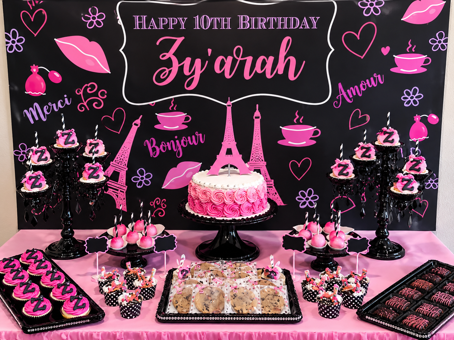 A dessert table with pink and black themed treats and a backdrop that says 'Happy 10th Birthday Z'yarah'. The table has cupcakes, cookies, cake pops, and a central cake with Eiffel Tower decorations, all in pink, black, and white. The backdrop featur