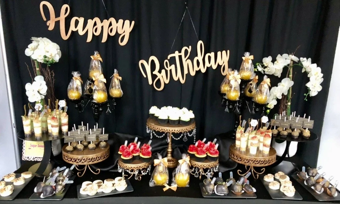 Birthday dessert table with cupcakes, cake pops, and other sweets, decorated with white orchids and a black backdrop with gold 'Happy Birthday' sign.