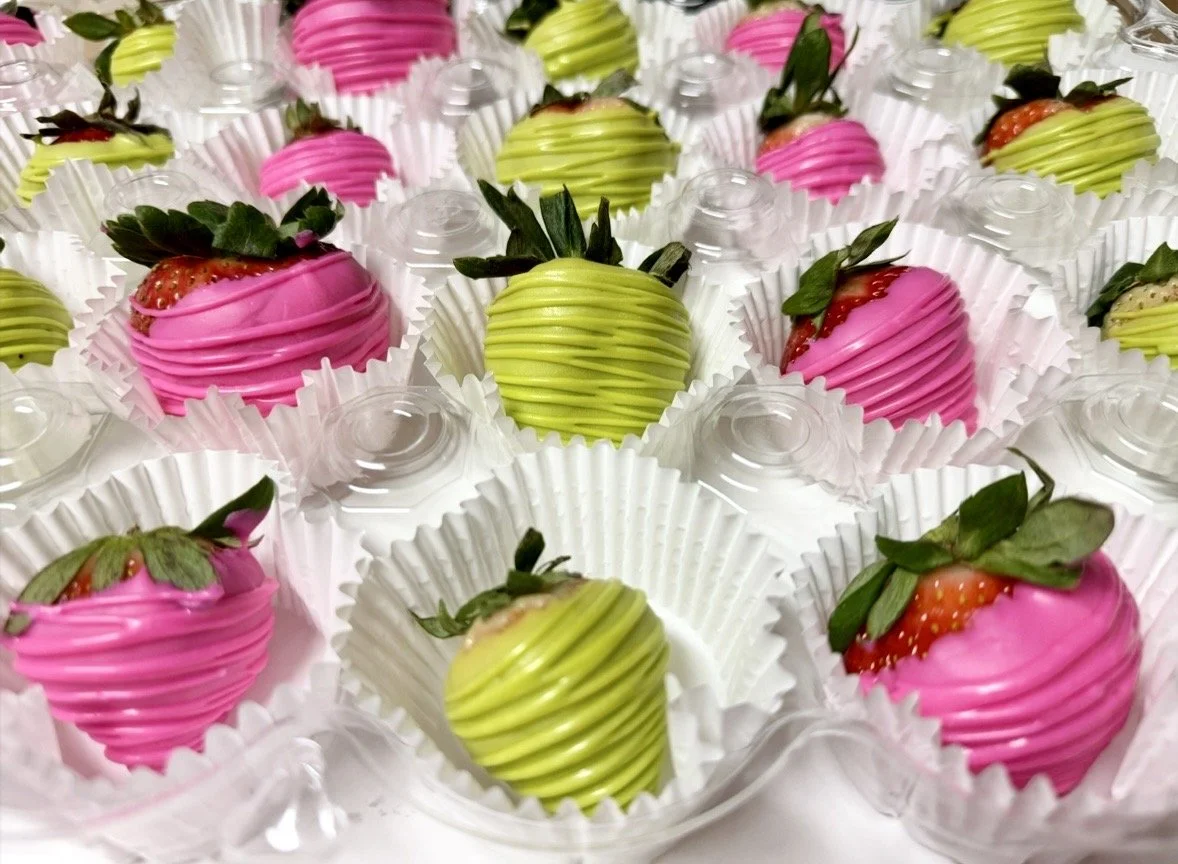 Strawberries dipped in pink and green chocolate with decorative drizzle, arranged in individual paper cups.