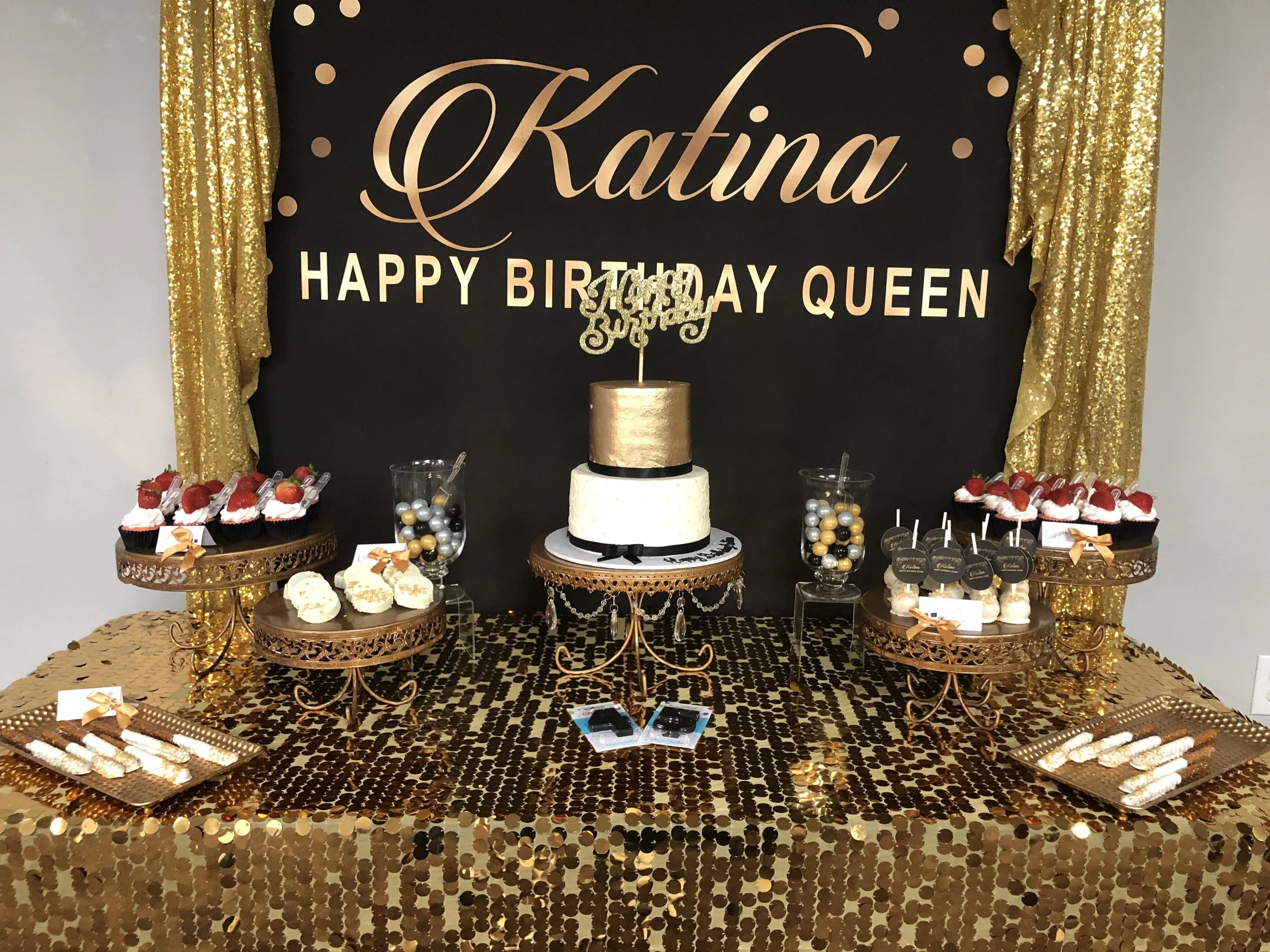 Table decorated for a birthday celebration with a black and gold theme. A large black backdrop with gold lettering that says 'Kafina' and 'Happy Birthday Queen,' flanked by gold sequin curtains. The table has gold chargers, cake and cupcake displays,