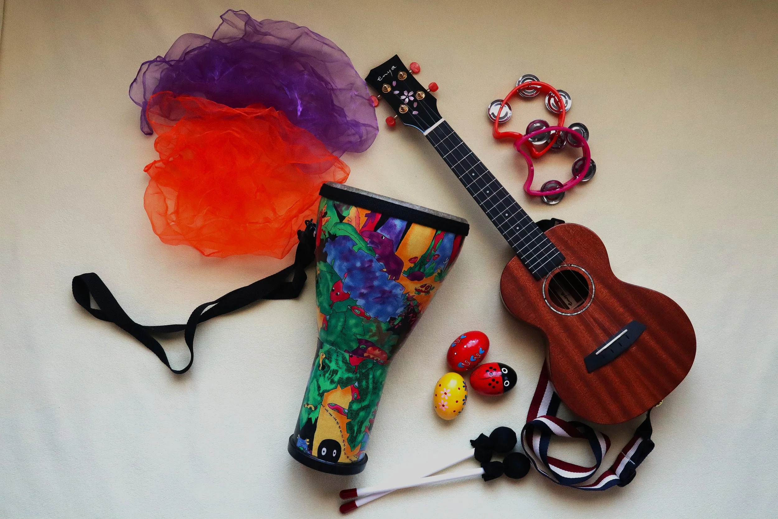 A collection of colorful children's toys and accessories on a white surface, including a small ukulele, a pair of heart-shaped tambourines, a decorated cup, several plastic Easter eggs, and some hair accessories.