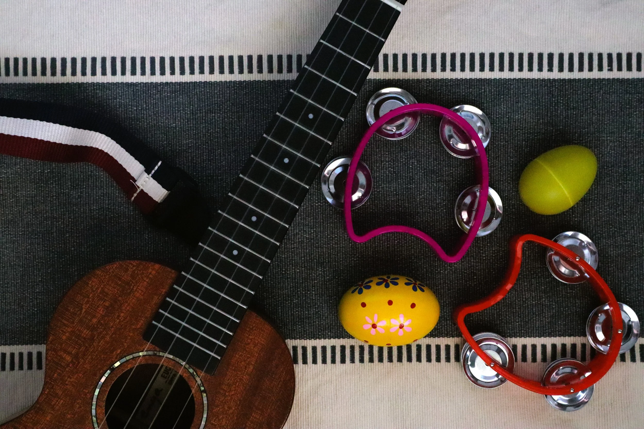 A collection of small musical and decorative objects on a striped fabric surface. Includes a small ukulele, several metal bottle caps, a pink heart-shaped wire frame, a yellow oval object with pink and blue floral designs, and other colorful items.