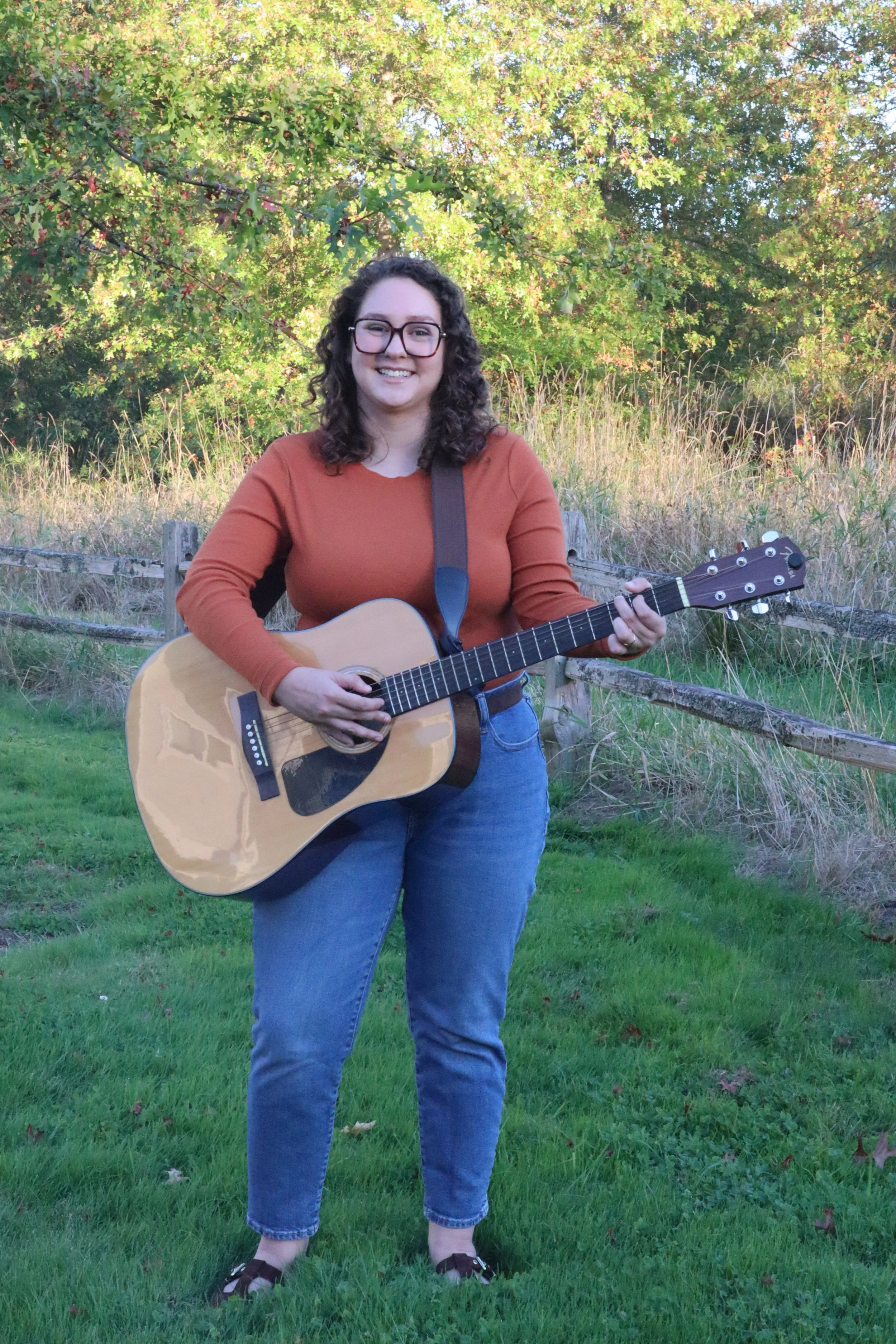 A woman with curly dark hair and glasses, wearing an orange long-sleeve shirt and blue jeans, playing an acoustic guitar outdoors in a grassy area with trees and a wooden fence in the background.