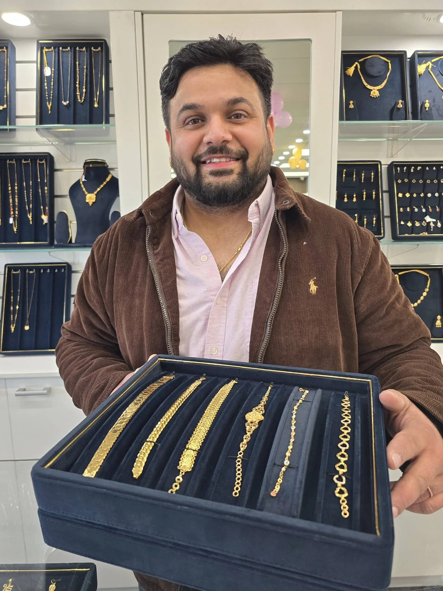 A man displaying a box of gold jewelry, including necklaces and bracelets, in a jewelry store with jewelry displays in the background.