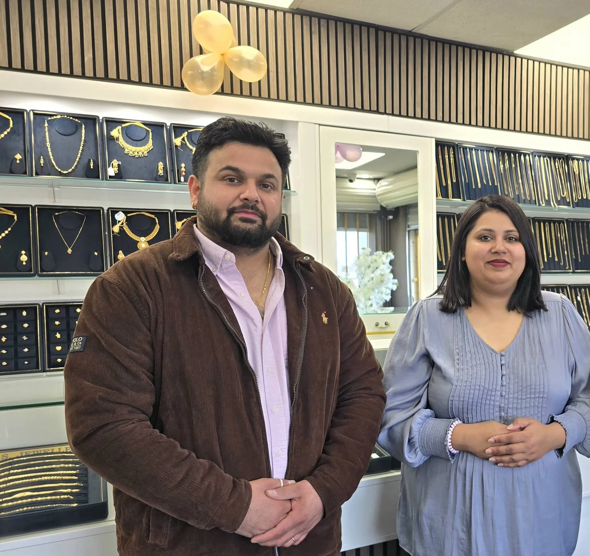 Two people are standing in front of a jewelry display. The man on the left has short dark hair, a beard, and is wearing a brown jacket and a pink shirt. The woman on the right has long dark hair and is wearing a light blue shirt. The jewelry display behind them features necklaces, earrings, and other jewelry pieces, with golden accents. The background includes a ceiling with wooden slats and a decorative balloon arrangement.
