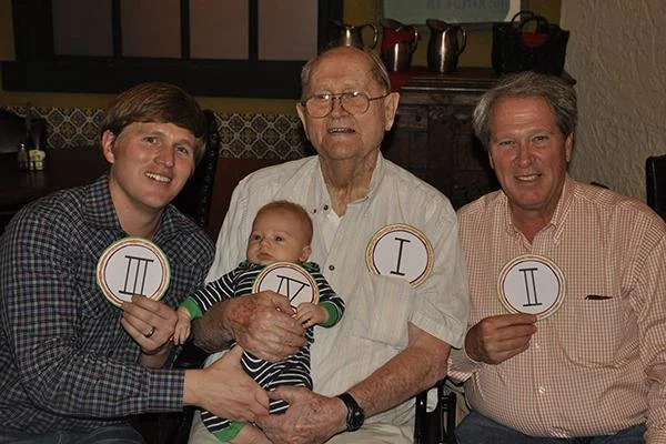 Four men, including a baby, sitting together and holding circular signs with Roman numerals I, II, and III.