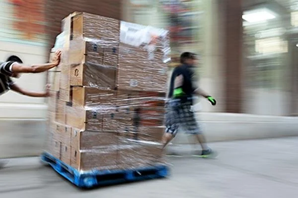 Two people are pushing a large pallet of cardboard boxes wrapped in plastic along a city sidewalk.