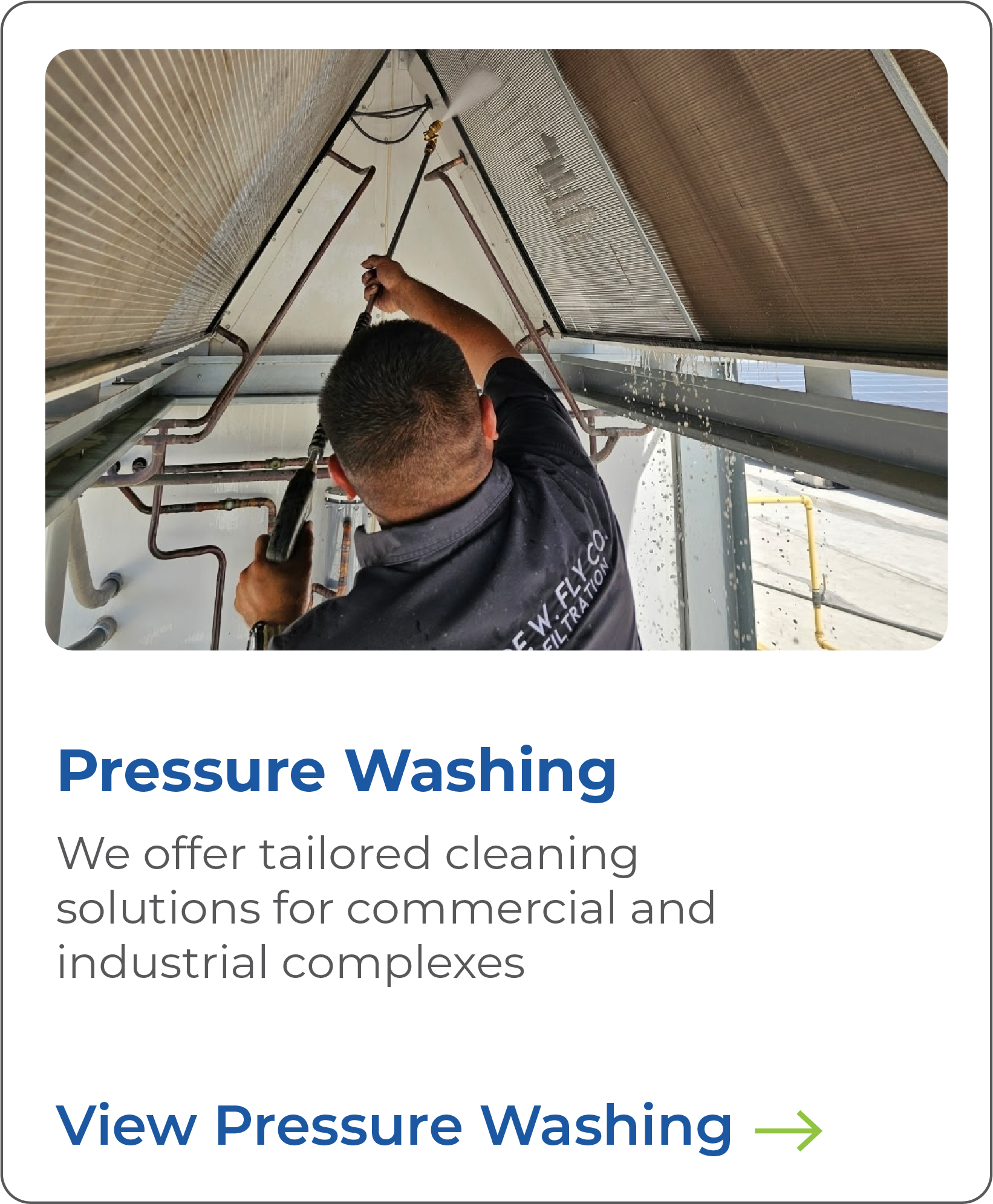 A worker cleaning the underside of an industrial air conditioning unit with a pressure washer. The worker is holding a spray gun connected to a hose, wearing a black shirt, with the back of his head visible. The unit has metal fins, and water can be seen spraying out.