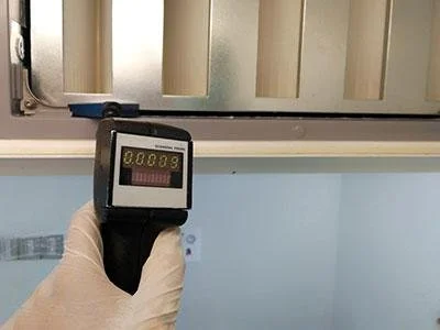 A hand holding a detector with a digital display showing a reading of 0.0005, in front of a wall with ridges.