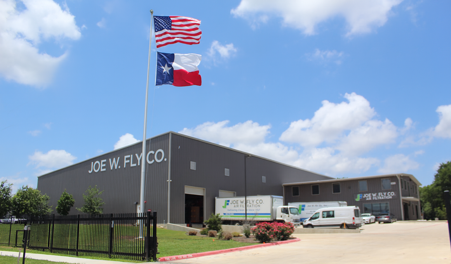 Exterior view of Joe W. Fly Co. building with American and Texas flags flying high on flagpoles, parked trucks and vans in front, green grass, and blue sky with clouds.