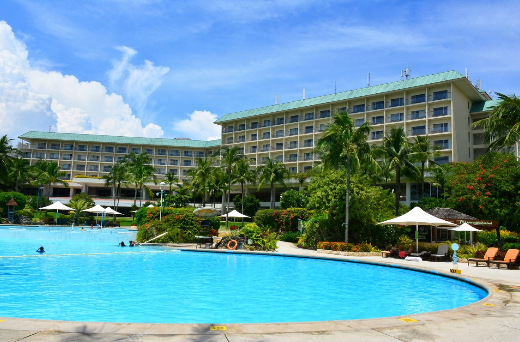 A Hilton hotel with a swimming pool in the foreground, surrounded by palm trees and lounge chairs, under a partly cloudy sky.