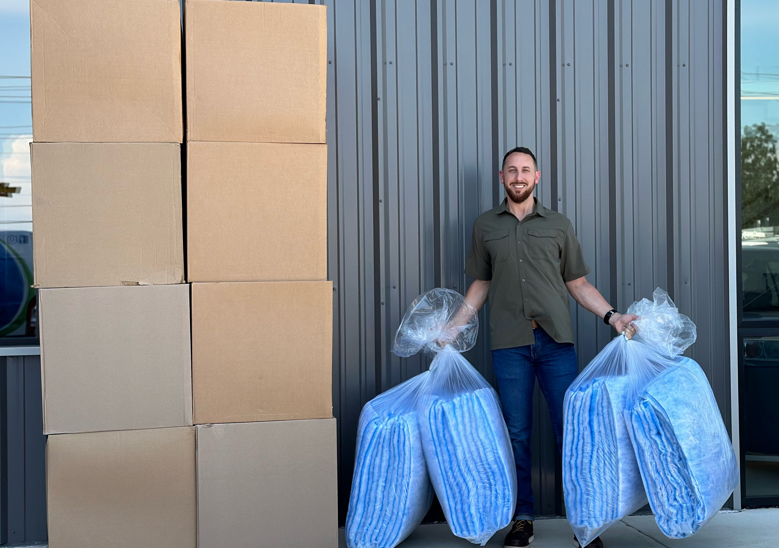 Smiling man standing outside a metal building with a wall behind him, holding 4 plastic filters bags, standing next to 8 large filter case boxes.