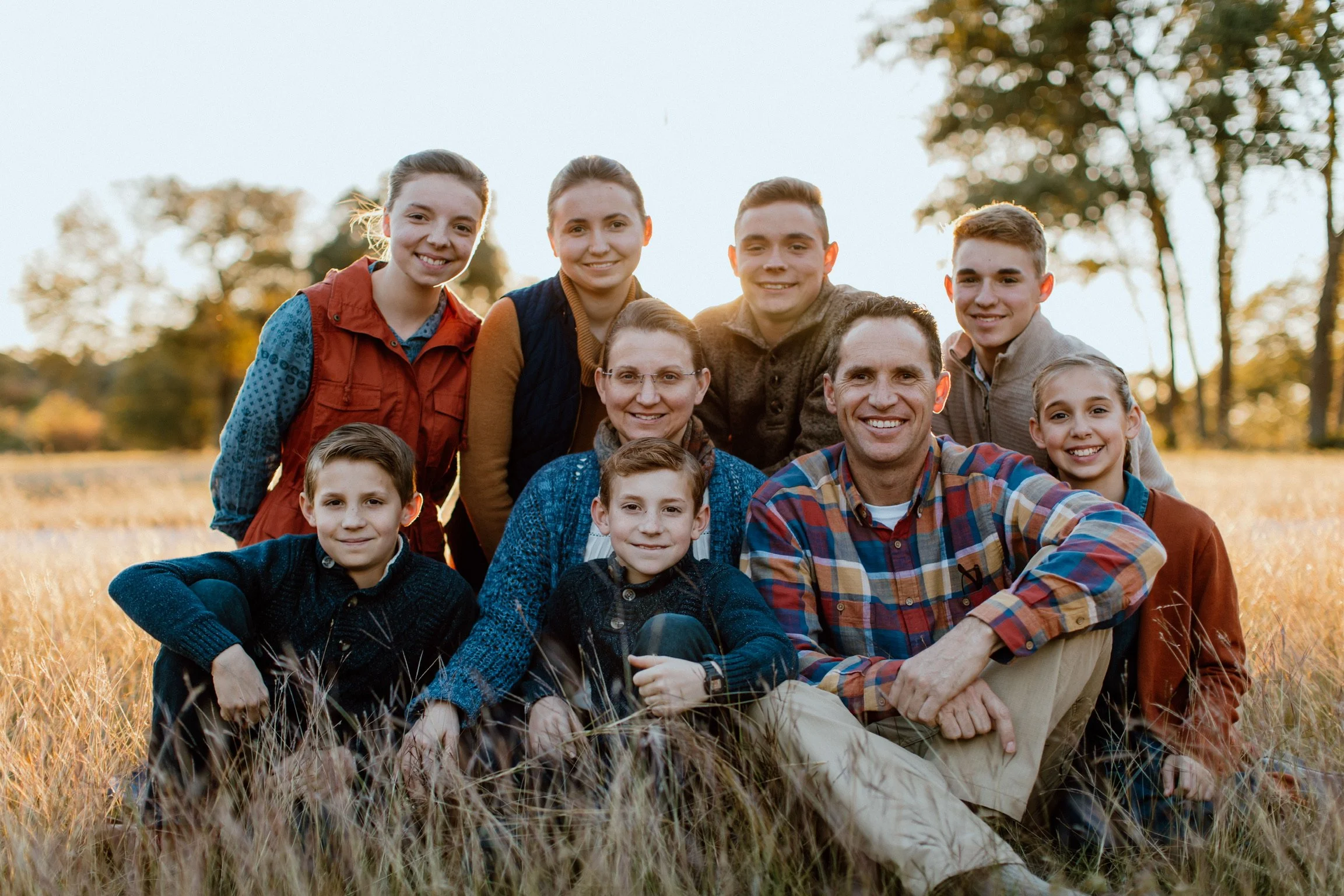 Family of ten people, including children and adults, posing outdoors in a field during sunset or late afternoon, smiling for a group photo.