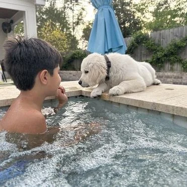 A young boy in a swimming pool face-to-face with a puppy lying on the pool's edge.