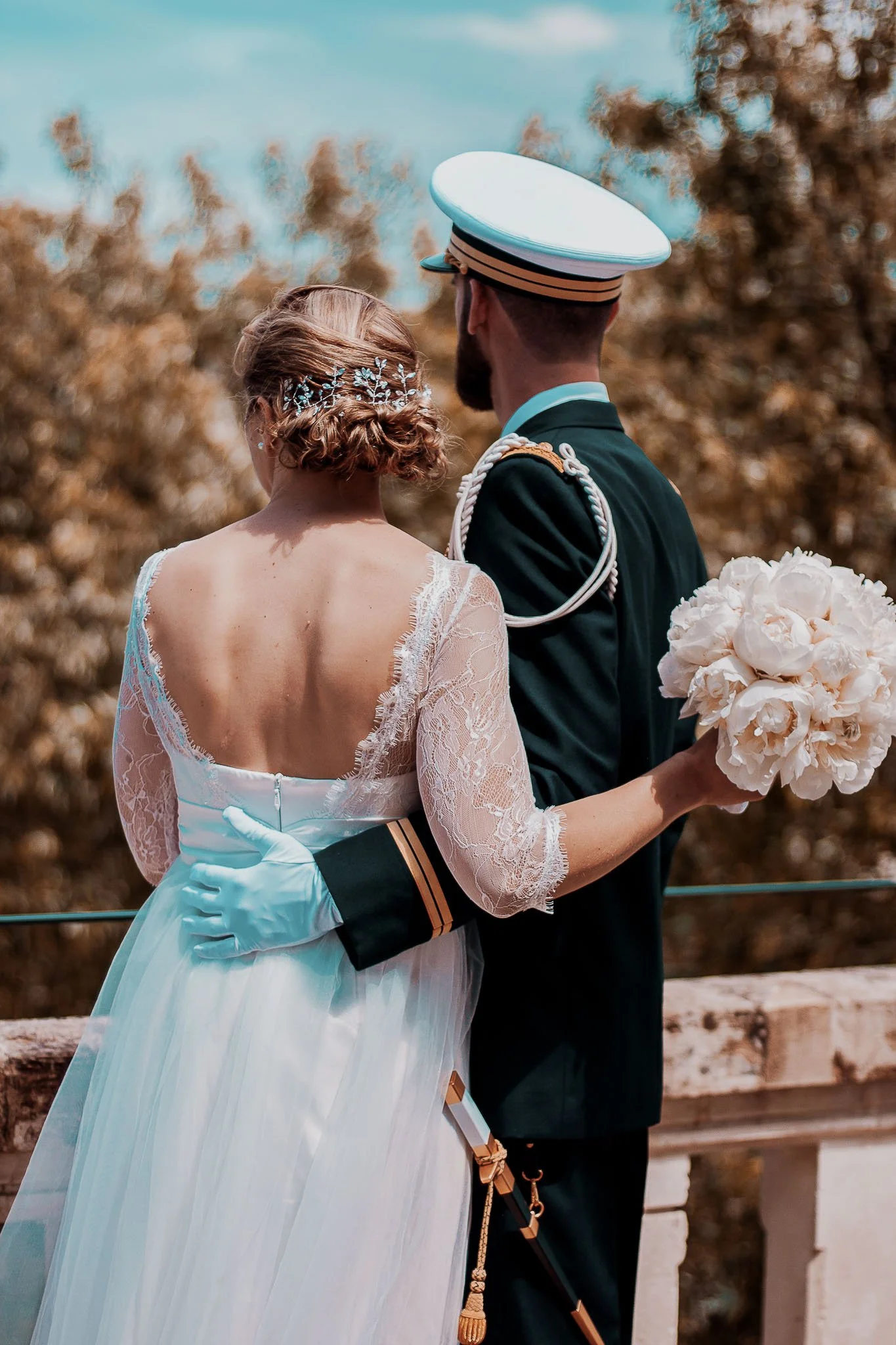 Un couple en tenue de mariage, l'homme en uniforme militaire, la femme en robe de mariée, tenant un bouquet de fleurs, vue de dos, dans un cadre naturel.