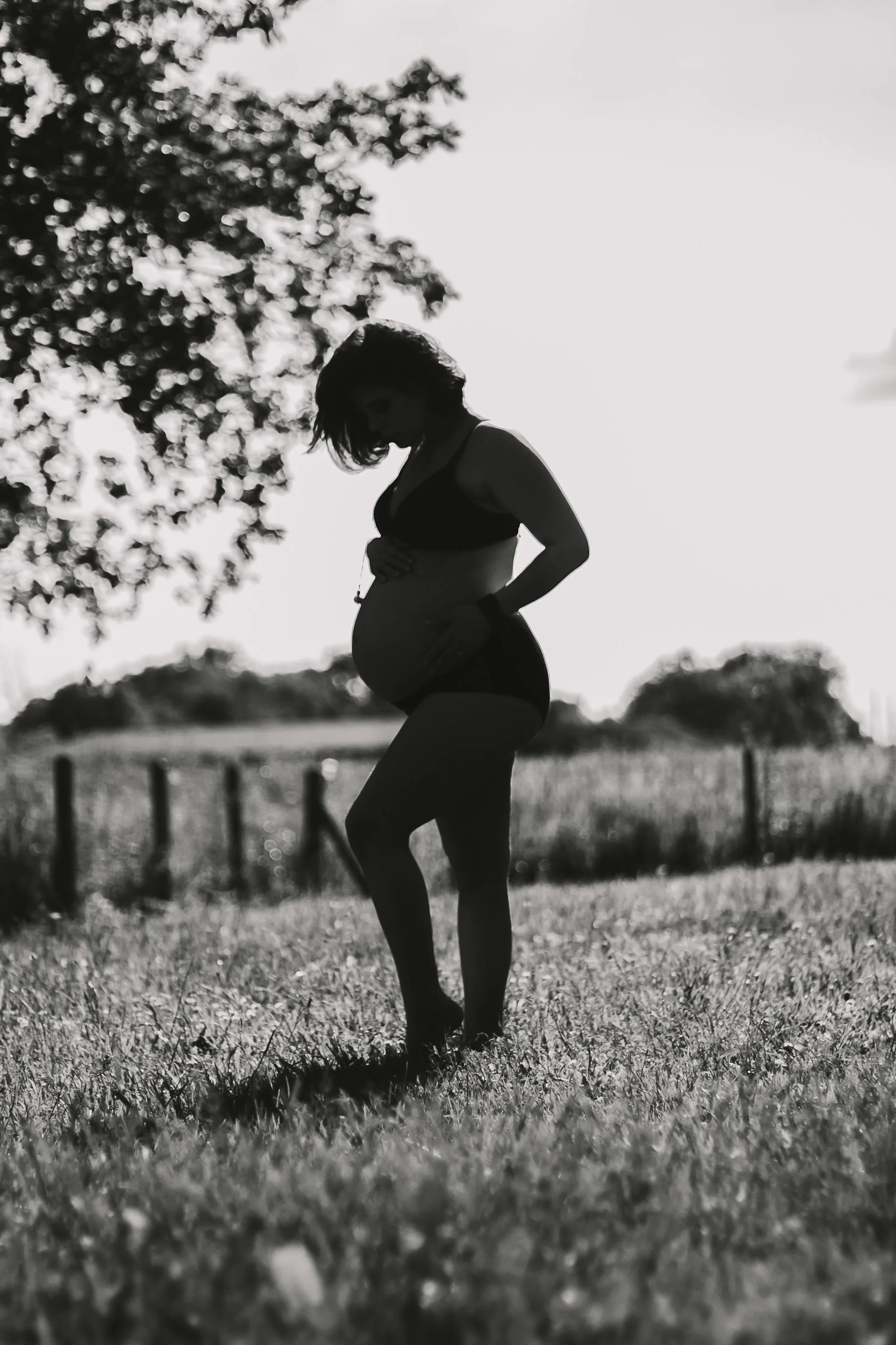 Silhouette d'une femme enceinte debout dans un champ, observant son ventre, avec des arbres en arrière-plan en noir et blanc.
