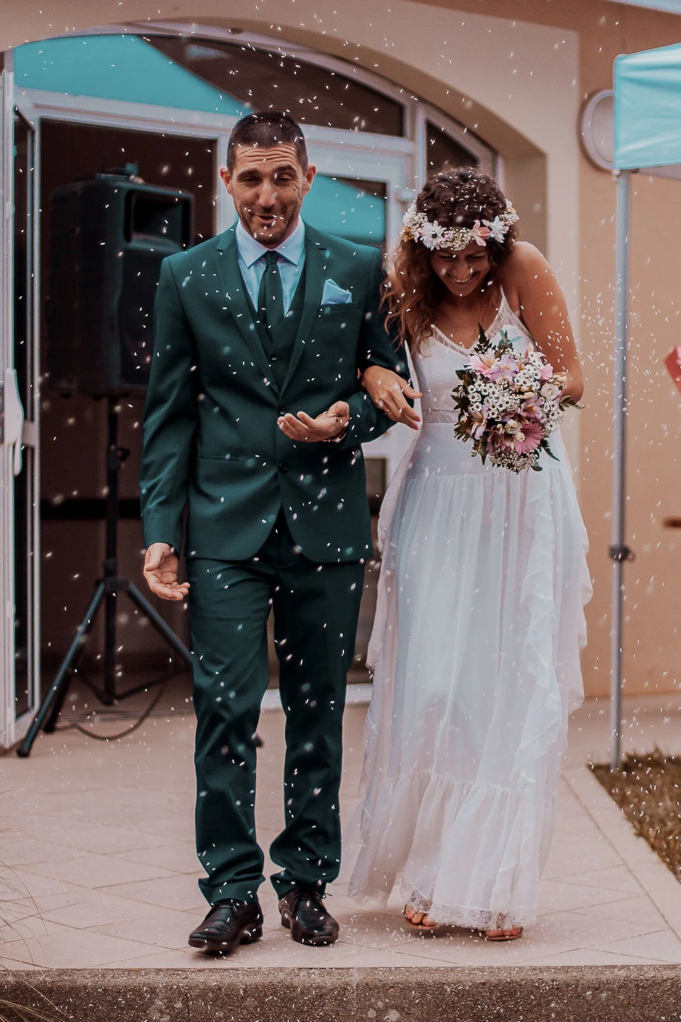Des mariés marchant ensemble lors d'une cérémonie, la femme portant une couronne de fleurs et tenant un bouquet, la scène étant décorée avec des confettis dans l'air.