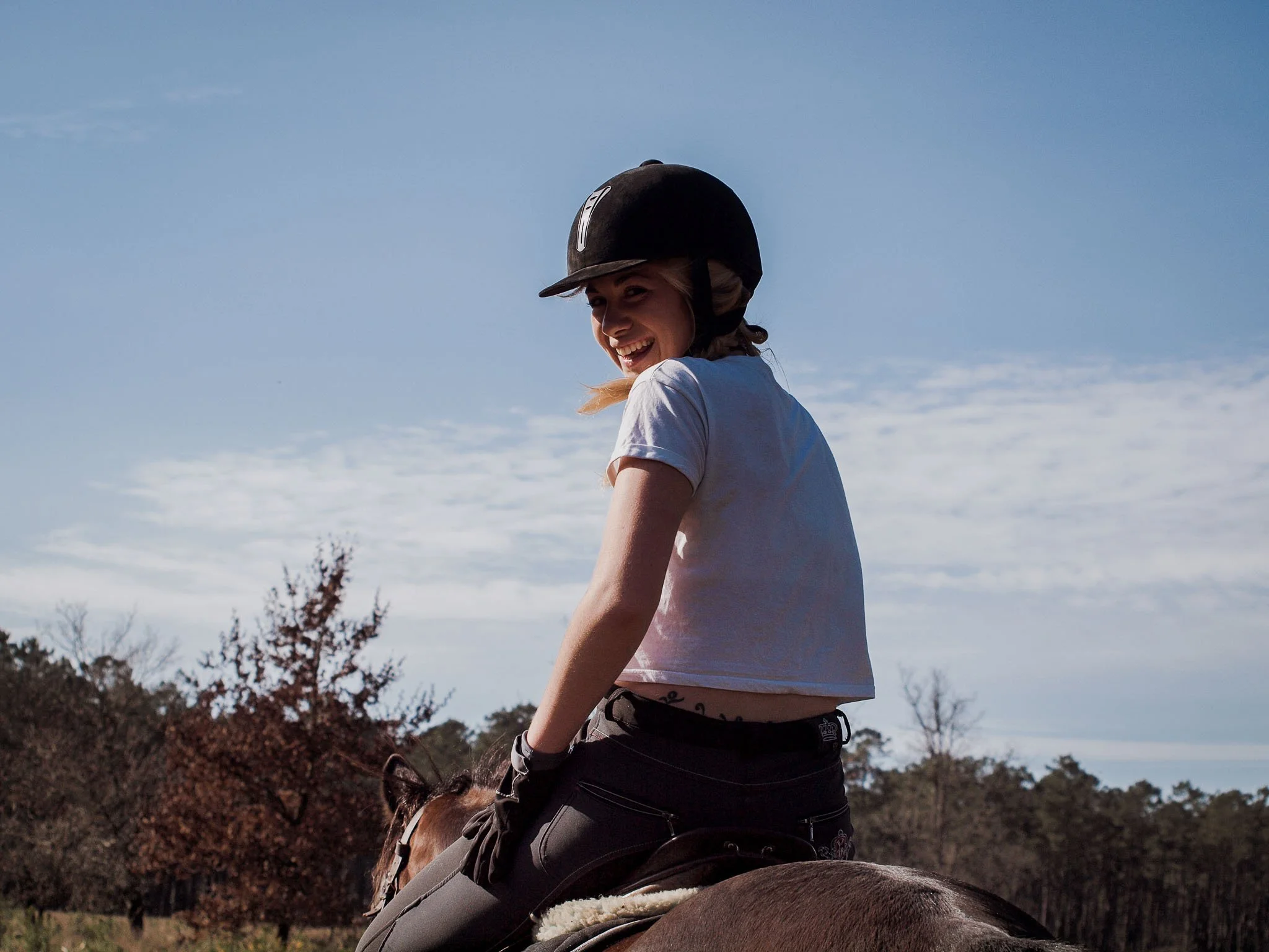 Femme souriante portant un casque d'équitation, montée à cheval dans un paysage naturel avec ciel bleu et arbres en arrière-plan.