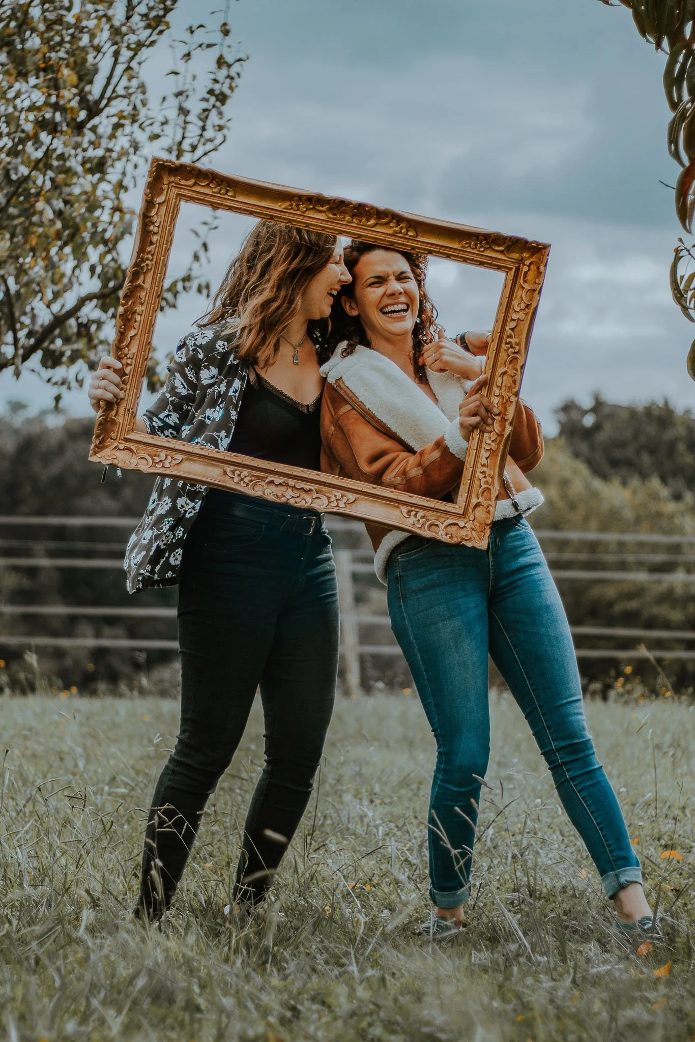 Deux femmes rient et posent avec un cadre en bois dans un champ en plein air, sous un ciel nuageux.