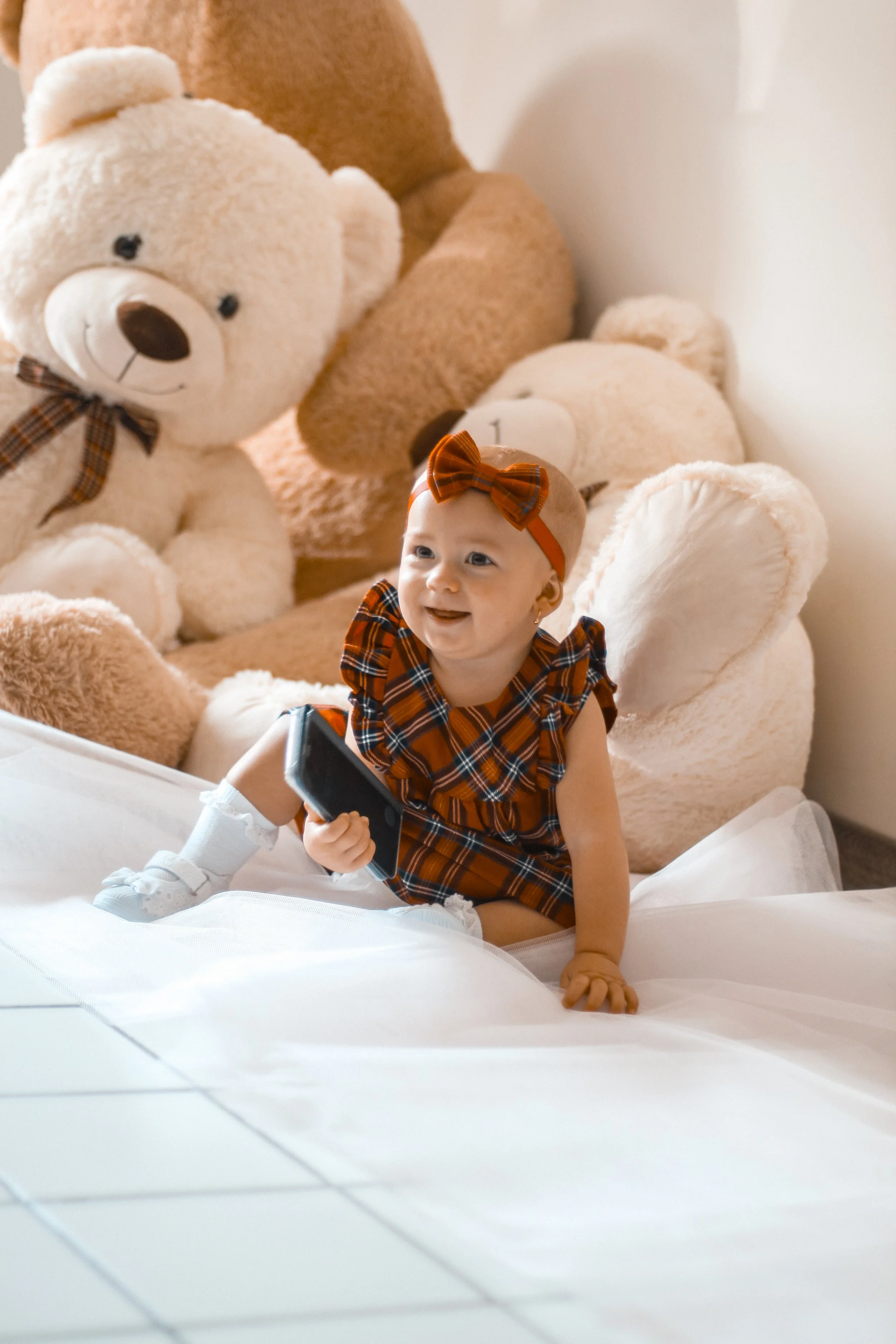 Un bébé habillé d'une robe tartan orange et noire, portant un bandeau coordonné, assis sur un lit blanc, tenant un téléphone portable, avec des ours en peluche en arrière-plan.
