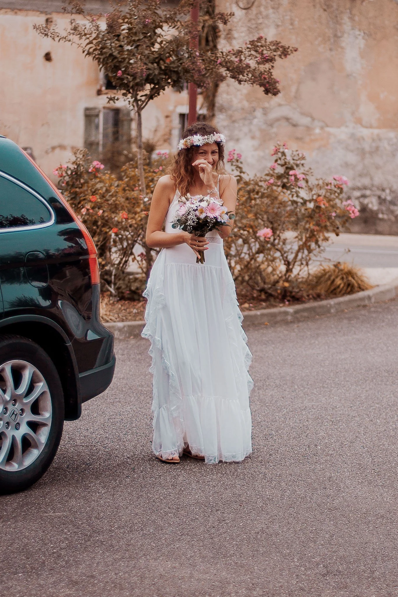 Une femme habillée d'une robe longue blanche, portant une couronne de fleurs sur la tête, tenant un bouquet de fleurs, debout sur une route en ville avec des plantes et un arbre à l'arrière-plan, et à côté d'une voiture noire.