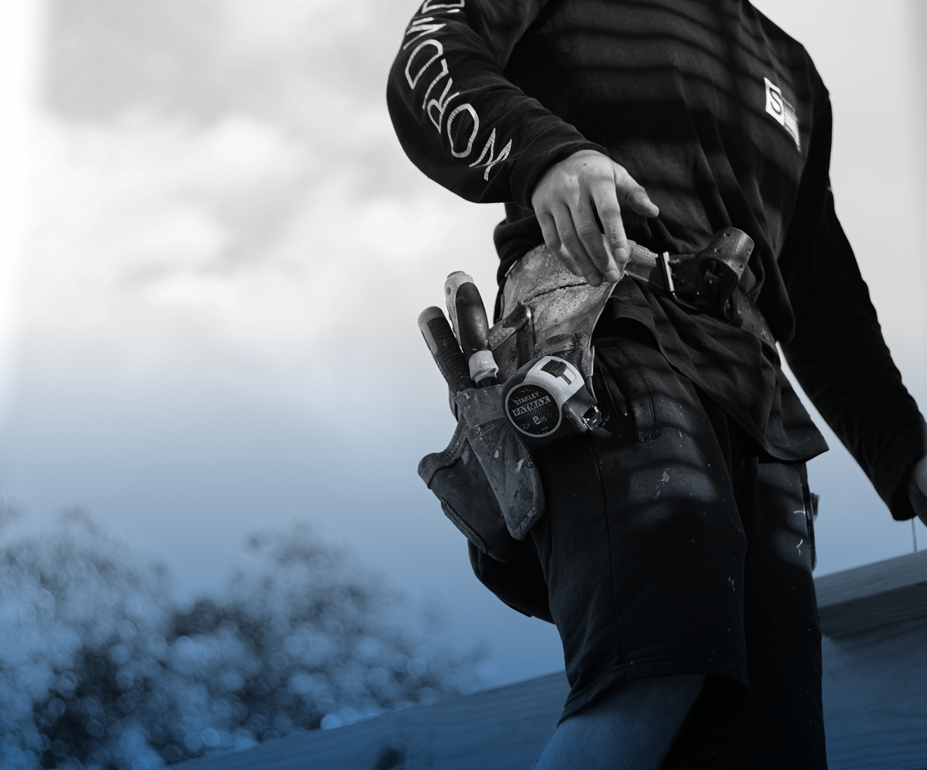A construction worker wearing black clothing and a tool belt with various tools, standing outdoors against a cloudy sky.
