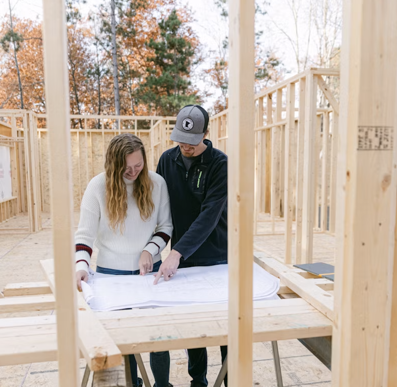 Two people, a man and a woman, reviewing building plans at a construction site for a wooden framing structure with trees in the background.