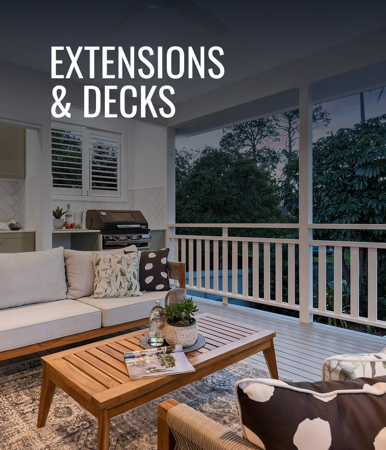 An outdoor living space with a covered deck, white railing, seating area with a sofa and chair, wooden coffee table with a plant and magazine, and a built-in grill in the background. Trees and evening sky visible beyond the deck.