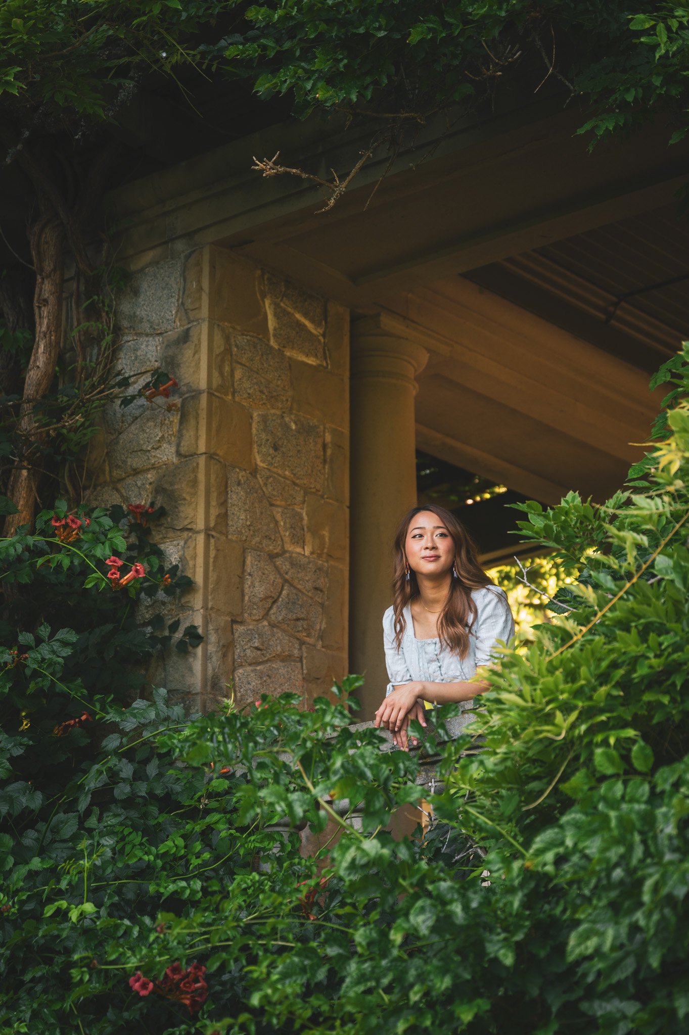 A woman with long brown hair wearing a white blouse standing amidst green plants and flowers outside a stone building with columns and a wooden roof.