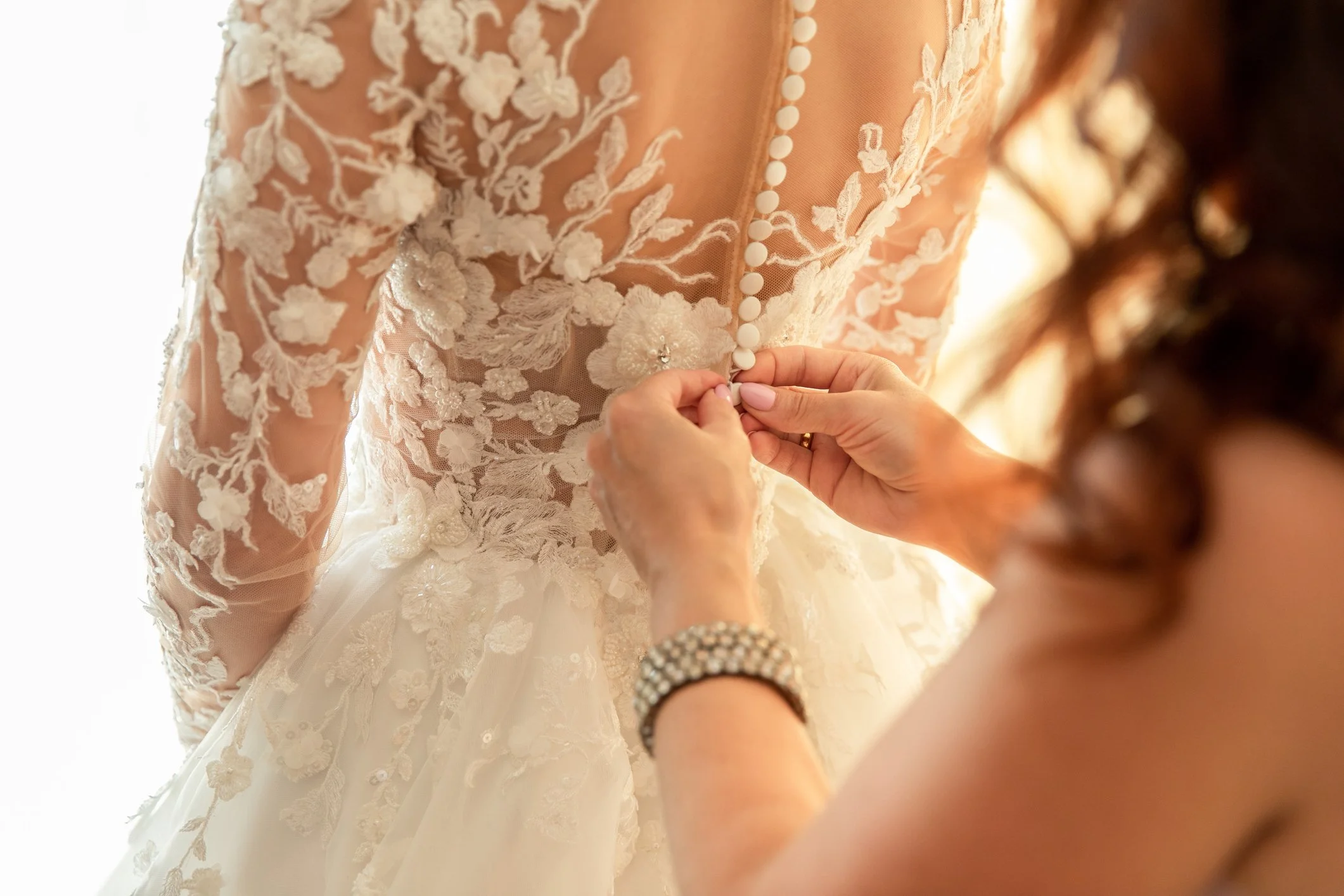 A bride is buttoning up her wedding dress, which features intricate lace embroidery with floral patterns. The dress has a row of small buttons down the back, and the bride's face is not visible in the image.