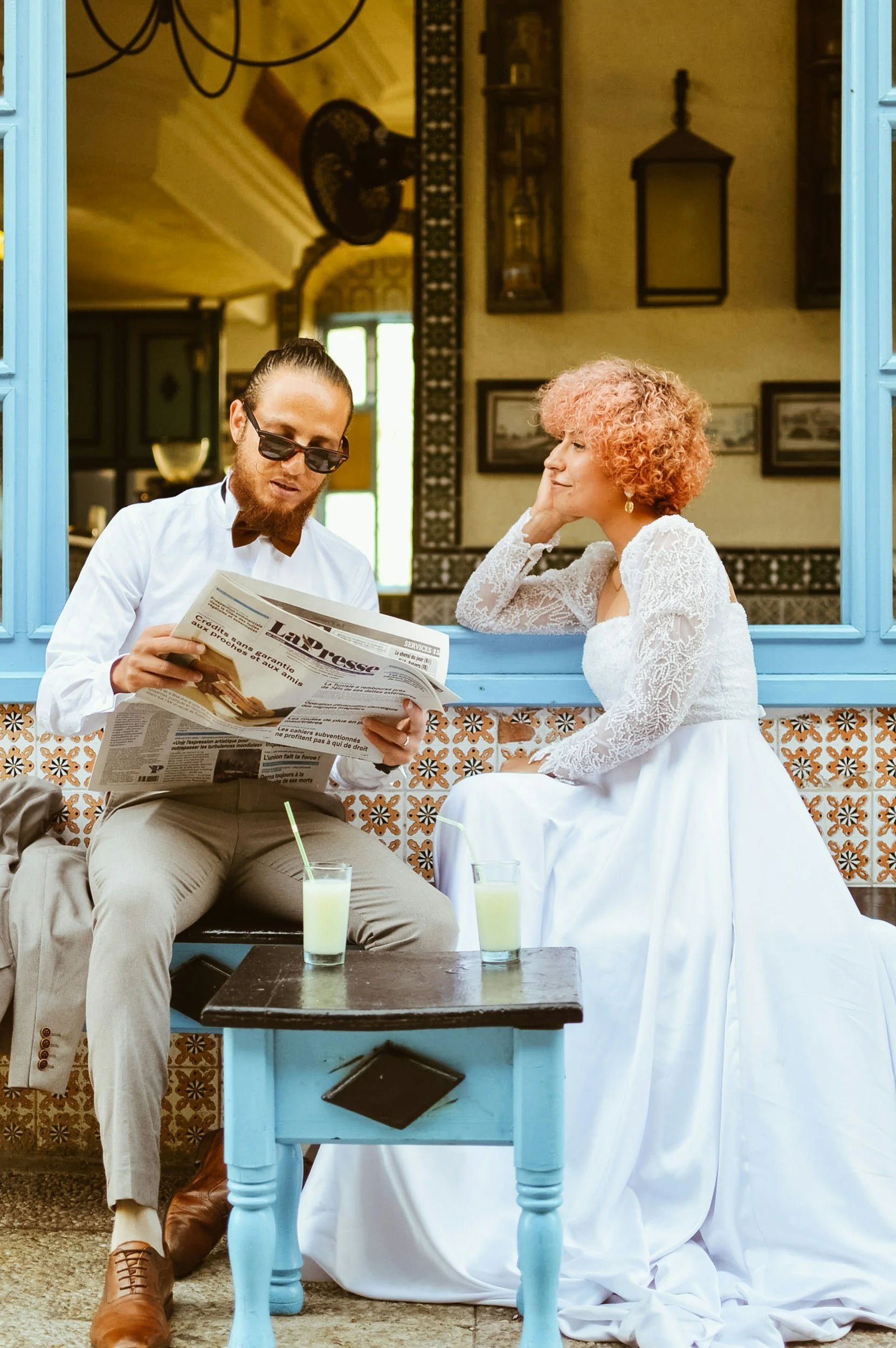 Man wearing sunglasses and a bow tie, reading a newspaper, sitting at a blue table with two drinks, and a woman in a white dress with curly hair sitting across from him inside a decorated room with a patterned wall, framed pictures, and a window.