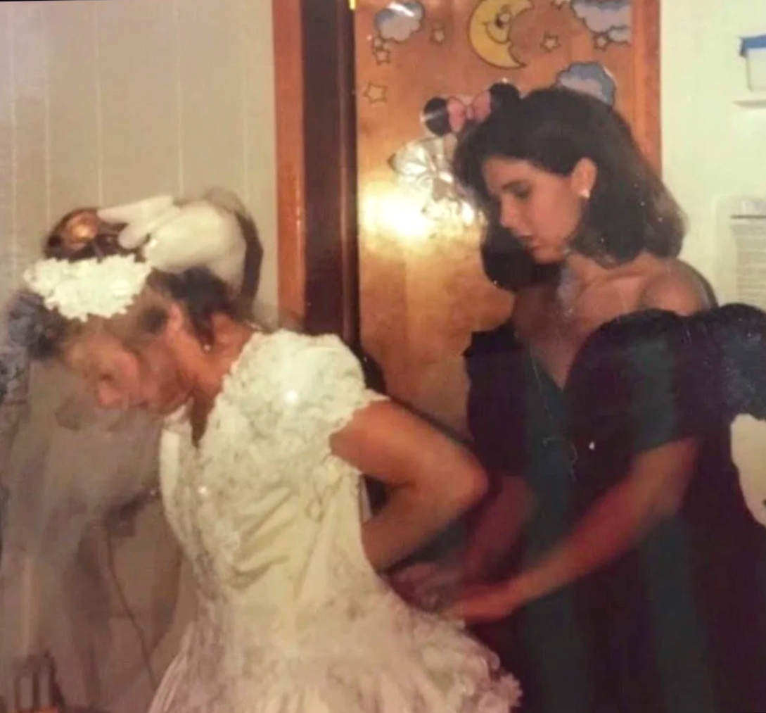 A bride in a white wedding dress and floral headpiece stands with her hands on her hips, while a woman in a black dress is helping her with the dress. The scene is indoors against a wooden door with a colorful moon and stars poster.
